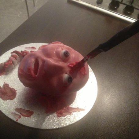 A decorated cake in the shape of a decapitated head with an exposed spine and a kitchen knife in it's forehead